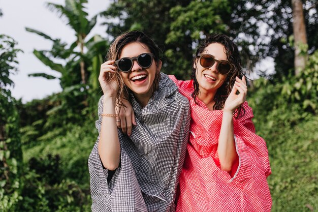 Alegres mujeres de cabello oscuro con gafas de sol riendo de la naturaleza. Turistas de buen humor con impermeables divirtiéndose en la jungla.