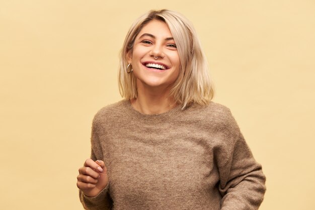 Alegría, felicidad y emociones positivas. Retrato de mujer joven hermosa con estilo en suéter acogedor divirtiéndose, riéndose de un buen chiste, sintiéndose feliz y despreocupado, posando aislado en la pared en blanco del espacio de la copia