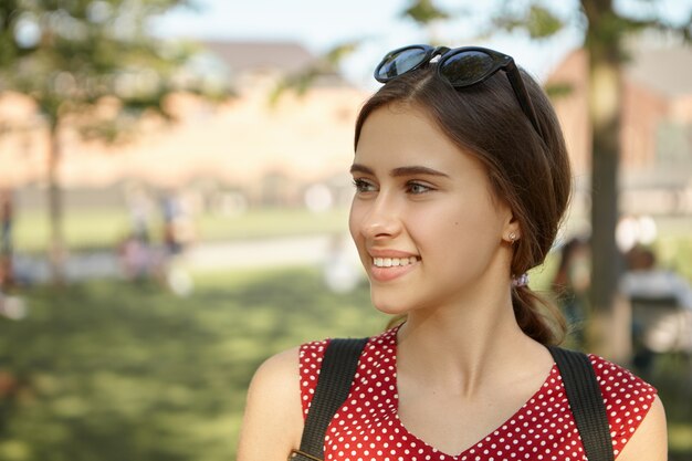 Alegría, felicidad, verano, vacaciones y concepto de viaje. Mujer joven atractiva alegre con gafas de sol en la cabeza y mochila sonriendo alegremente, emocionado mientras hace turismo en alguna ciudad europea