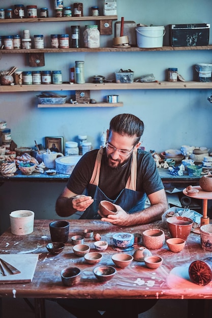 Un alfarero pensativo y entusiasta con gafas en su propio taller está trabajando en una nueva tetera hecha a mano.