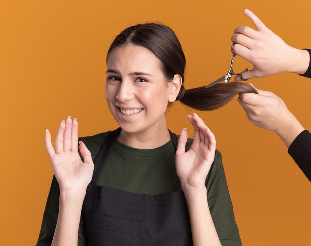 Alguien corta la trenza con unas tijeras para adelgazar el cabello de una joven barbero morena molesta en uniforme de pie con las manos levantadas aisladas en la pared naranja con espacio de copia