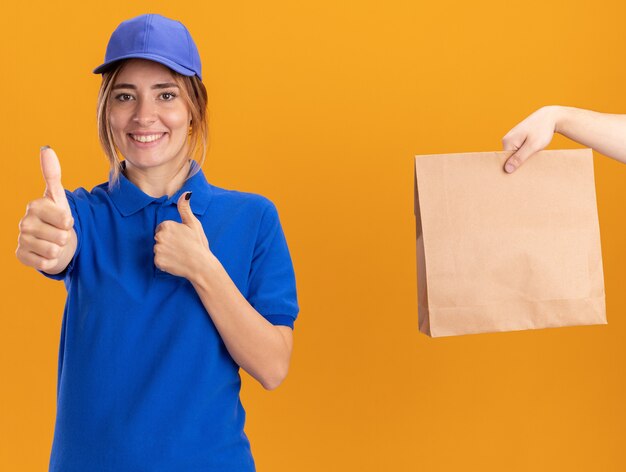 Alguien le da un paquete de papel a la sonriente joven repartidora bonita en uniforme, pulgar hacia arriba con las dos manos en naranja