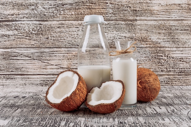 Algunas botellas de leche con dividido en medio coco en el fondo de madera blanco, vista lateral.