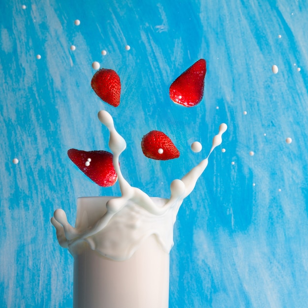 Algunas fresas que caen en la leche en el fondo texturizado azul ciánico, vista lateral.