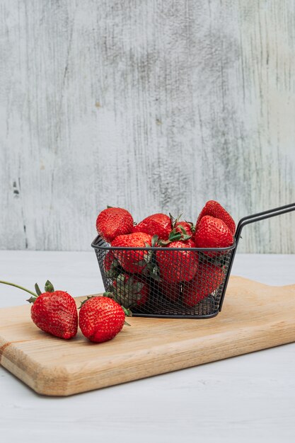 Algunas fresas con varias fresas a su alrededor en una canasta negra sobre tabla para cortar madera y fondo blanco, vista lateral