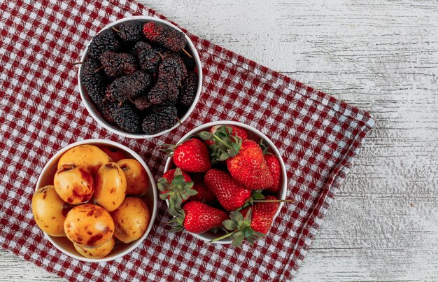 Algunas moras con fresas, nísperos en cuencos sobre tela y fondo de madera clara, vista superior.