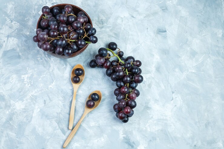 Foto gratuito algunas uvas negras en cuenco de arcilla y cucharas de madera sobre fondo gris sucio, vista superior.