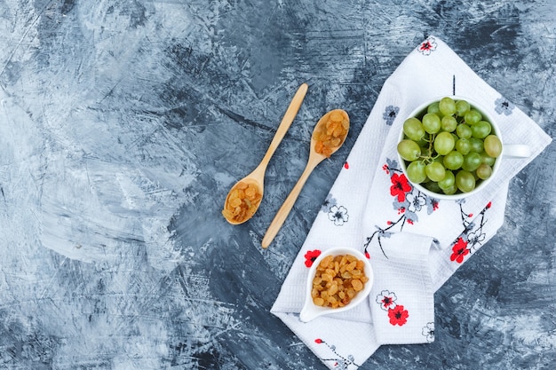 Algunas uvas verdes con pasas en una taza blanca sobre yeso sucio y fondo de toalla de cocina, vista superior.