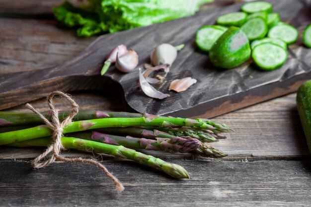 Algunos espárragos con rodajas de pepino, ajo, lechuga en una tabla de cortar sobre fondo de madera oscura, vista de ángulo alto.