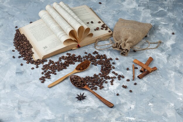 Algunos granos de café con café molido, libro, especias, saco en una cuchara de madera sobre fondo gris sucio, vista de ángulo alto.