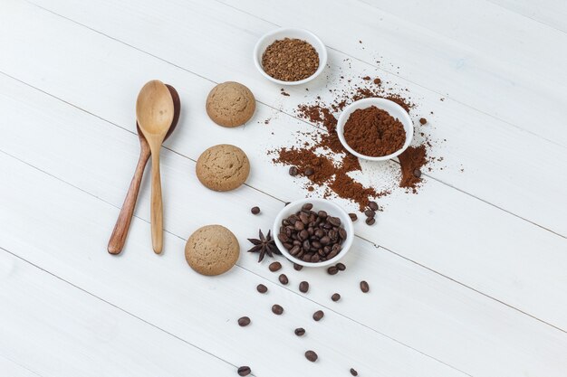 Algunos granos de café con galletas, cucharas de madera, café molido en un recipiente sobre fondo de madera, vista de ángulo alto.