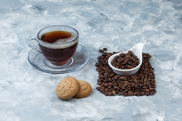 Algunos granos de café con galletas, taza de café en una jarra de porcelana blanca sobre fondo de mármol azul, primer plano.