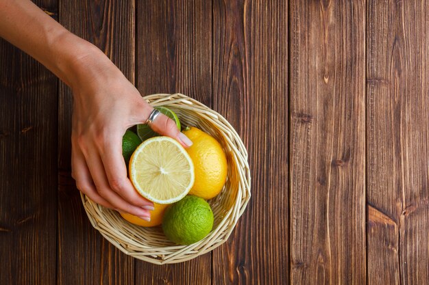 Algunos limones con mano sosteniendo la mitad de limón en una canasta sobre fondo de madera, vista superior.