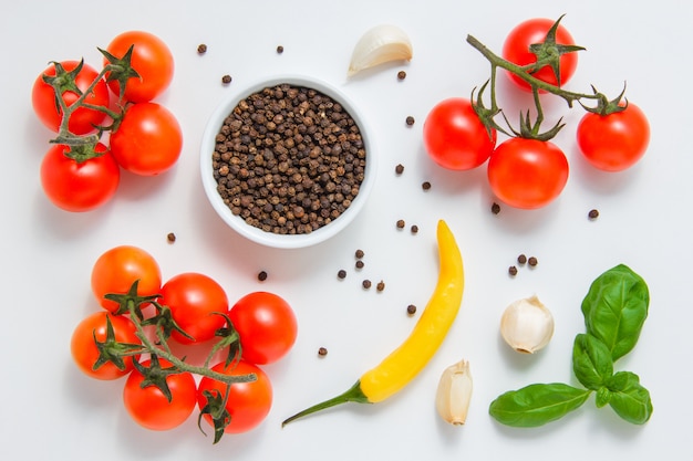 Algunos racimos de tomates con un tazón de pimienta negra, ajo, hojas, ají en la superficie blanca vista desde arriba.