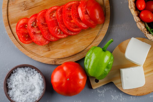 Algunos tomates en rodajas con queso, pimiento verde, sal en una tabla de cortar sobre una superficie gris, vista superior