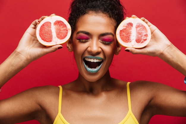 Alimentos moda encantada mujer afroamericana divirtiéndose sosteniendo dos mitades de pomelo maduro fresco en la cara, aislado sobre la pared roja