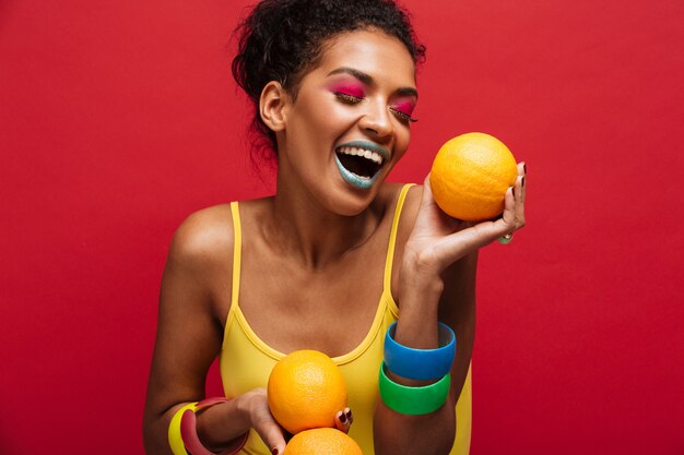 Alimentos moda mujer alegre de raza mixta con maquillaje colorido divirtiéndose con muchas naranjas maduras en las manos, aislado sobre la pared roja