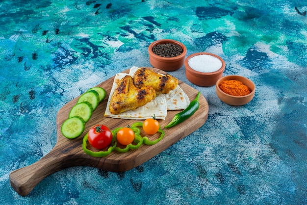 Alitas al horno, lavash y verduras sobre una tabla de cortar sobre la superficie azul