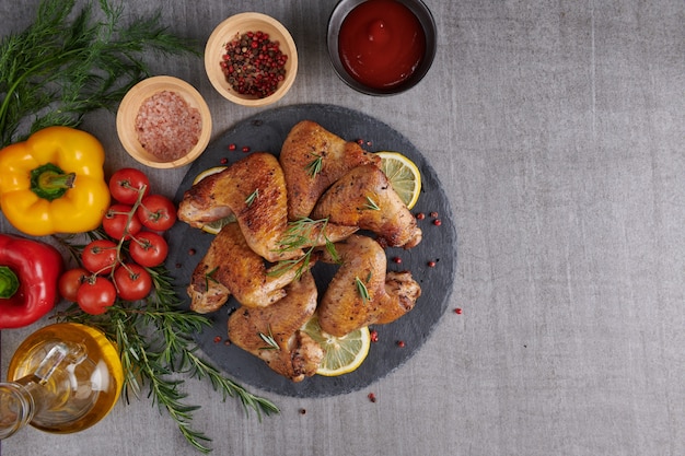 Alitas de pollo asadas en salsa barbacoa y ensalada mixta de verduras con pepitas de romero, sal en placa de piedra negra sobre mesa de piedra gris.