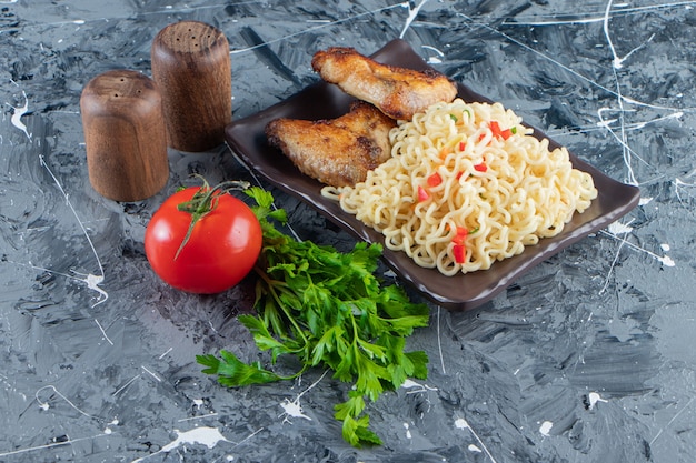 Alitas de pollo y fideos en un plato junto a verduras, sobre la superficie de mármol.