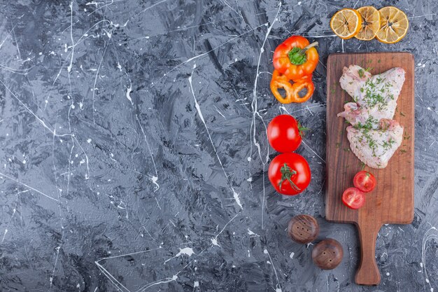 Alitas de pollo y tomates en rodajas sobre una tabla de cortar junto a una variedad de verduras en la superficie azul