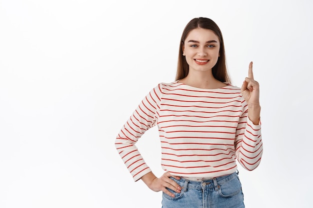 Allí arriba. Sonriente mujer atractiva que señala con el dedo en la parte superior, muestra el anuncio de copyspace hacia arriba, señala en la dirección, muestra el camino, se para sobre el fondo blanco.