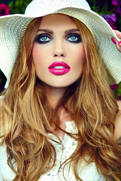 Alta moda look.glamor closeup retrato de hermosa sexy elegante rubia joven modelo con maquillaje brillante y labios rosados con piel limpia perfecta en sombrero cerca de flores de verano