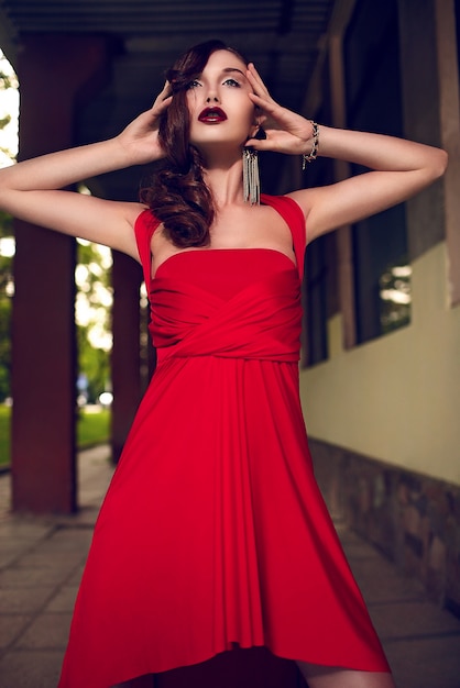 Alta moda look.glamor closeup retrato de hermosa sexy morena elegante modelo de mujer joven caucásica con maquillaje brillante, con labios rojos oscuros, en verano brillante vestido de noche rojo al aire libre