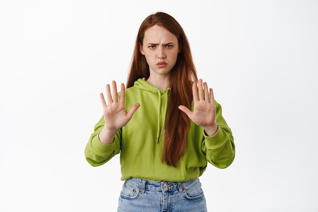 Alto ahí. Mujer pelirroja enojada y seria estira la mano para bloquear, rechazar la acción, prohibir y rechazar a la persona, disgustar y decir no, de pie sobre fondo blanco.