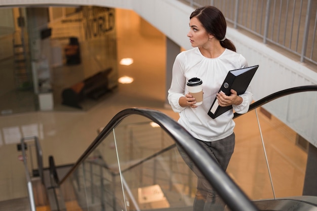 Alto ángulo de empresaria con carpeta y café en la escalera mecánica