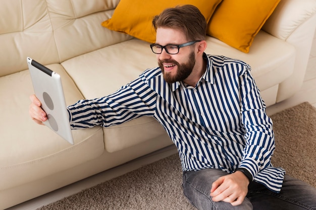 Alto ángulo de hombre sonriente en casa con tableta junto al sofá