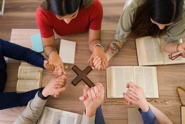 Alto ángulo de hombres y mujeres jóvenes católicos tomándose de la mano mientras rezan juntos alrededor de una mesa con una cruz cristiana