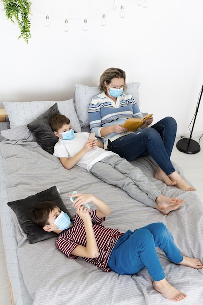 Alto ángulo de niños en la cama con máscaras médicas y madre leyendo un libro