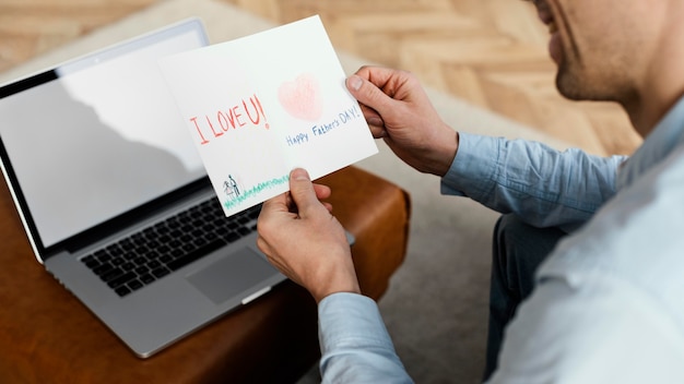 Foto gratuita alto ángulo de padre leyendo la tarjeta del día del padre mientras trabaja en la computadora portátil