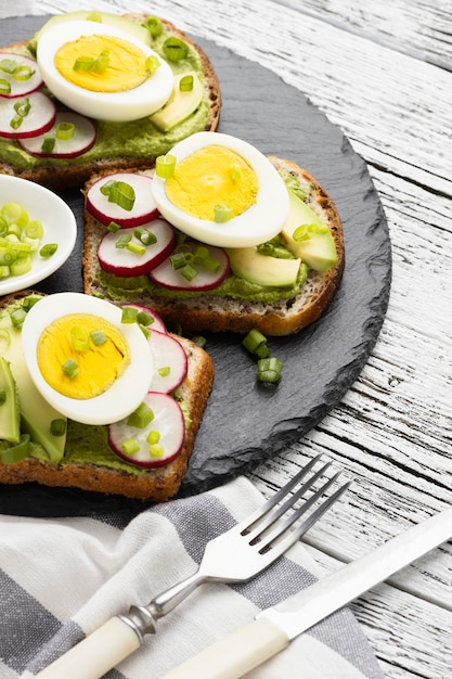 Alto ángulo de sándwiches de huevo y aguacate en pizarra con cubiertos