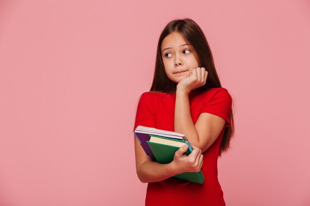 Alumna pensativa sosteniendo libros y mirando a un lado sobre rosa