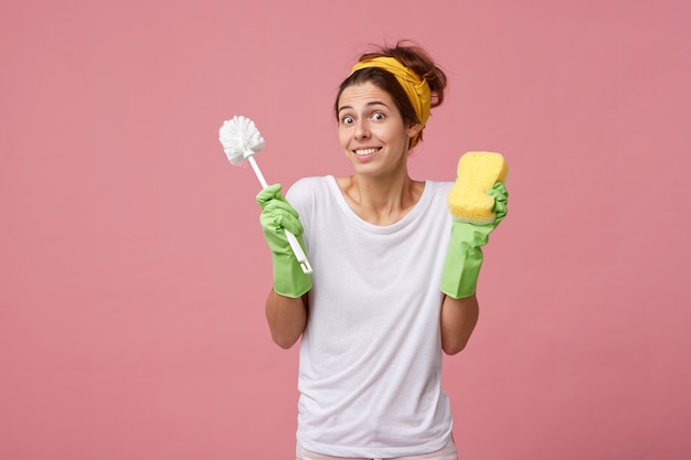 Ama de casa atractiva joven en ropa casual con guantes sosteniendo spray de lavado y cepillo con mirada de sorpresa encogiéndose de hombros mirando con desconcierto aislado sobre pared rosa