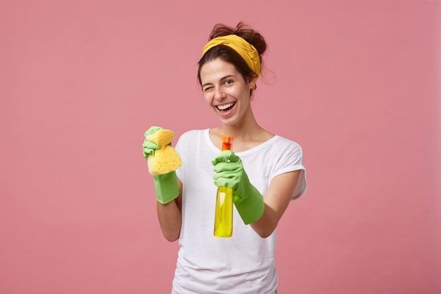 Foto gratuita ama de casa en camiseta blanca y guantes verdes con esponja y limpiador en las manos parpadeando con sus ojos con expresión alegre mientras se lava. joven bonita a mujer haciendo trabajo doméstico