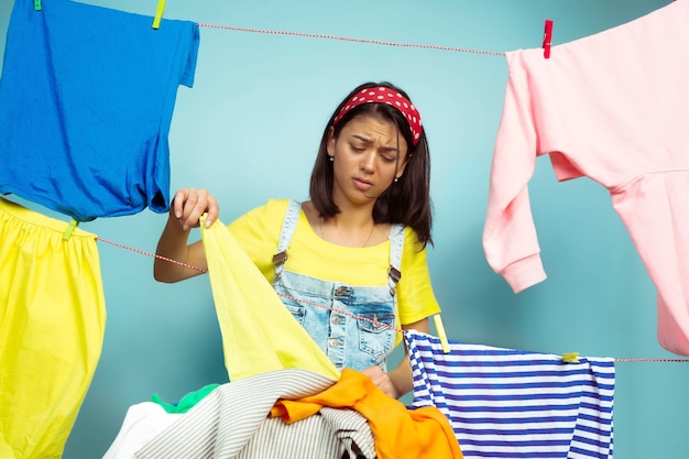 Ama de casa cansada y hermosa que hace las tareas domésticas aisladas sobre fondo azul. Mujer caucásica joven rodeada de ropa lavada. Vida doméstica, obras de arte brillantes, concepto de limpieza. Parece molesto.