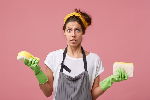 Ama de casa desconcertada con pañuelo amarillo en la cabeza, delantal, guantes de goma protectores sosteniendo dos esponjas ordenadas con mirada insatisfecha y sorprendida mientras va a limpiar su apartamento. Limpieza interna