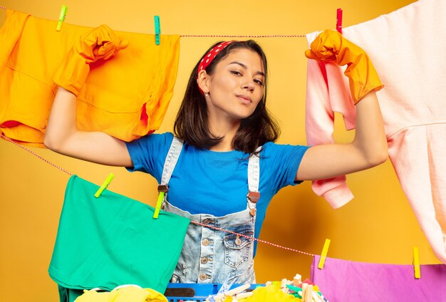 Ama de casa divertida y hermosa haciendo quehaceres domésticos aislados en el espacio amarillo. Joven mujer caucásica rodeada de ropa lavada