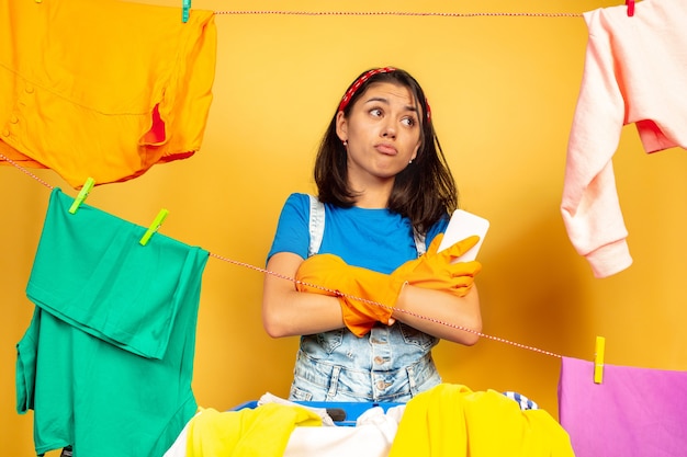 Ama de casa divertida y hermosa que hace las tareas domésticas aisladas sobre fondo amarillo. Mujer caucásica joven rodeada de ropa lavada. Vida doméstica, obras de arte brillantes, concepto de limpieza. Manos cruzadas.