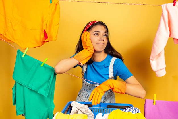 Ama de casa divertida y hermosa que hace las tareas domésticas aisladas sobre fondo amarillo. Mujer caucásica joven rodeada de ropa lavada. Vida doméstica, obras de arte brillantes, concepto de limpieza. Soñadora.