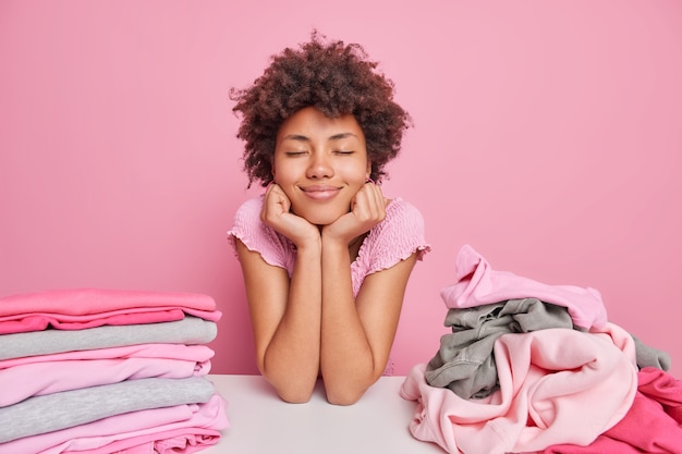 Ama de casa de ensueño complacida se inclina a la mesa con pilas de ropa limpia, mantiene las manos debajo de la barbilla, los ojos cerrados, toma un descanso después de doblar la ropa aislada sobre la pared rosa. Concepto de quehaceres domésticos