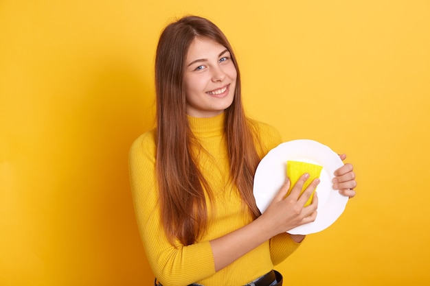 Ama de casa feliz demostrando el proceso de lavado, sosteniendo un plato blanco y una esponja en las manos y vistiendo ropa informal