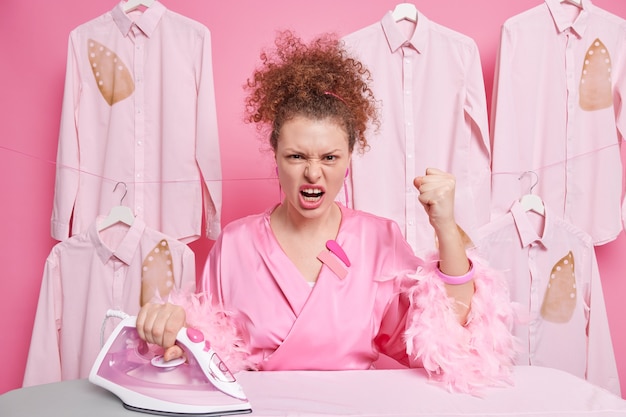 Ama de casa indignada molesta odio planchar aprieta los puños grita enojada tiene el pelo rizado vestido con ropa doméstica. Trabajador de lavandería irritado plancha camisas en el salón de tintorería. Concepto de limpieza