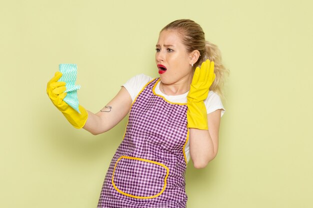 Ama de casa joven en camisa y capa púrpura guantes amarillos posando con expresión de disgusto en verde