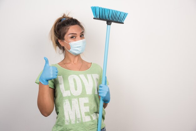 Ama de casa joven con mascarilla y escoba haciendo Thumbs up firmar en la pared blanca.