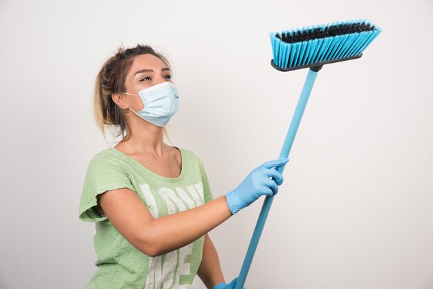 Ama de casa joven con mascarilla mirando escoba en la pared blanca.