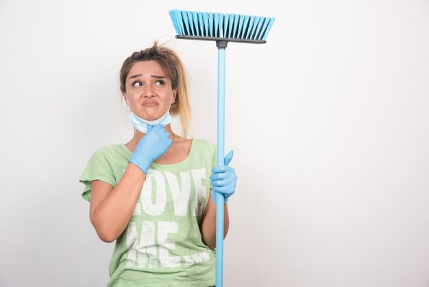 Ama de casa joven con mascarilla sosteniendo una escoba mientras mira hacia arriba confundida.